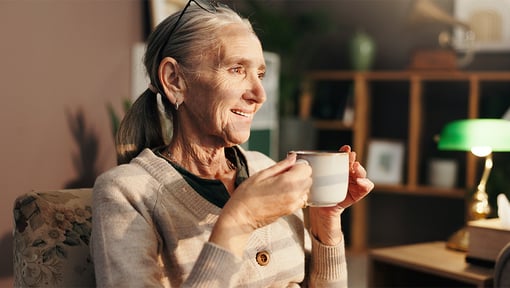 Older adult woman relaxing at home with a warm beverage, seated in a cozy living space with sunlight streaming through the window.