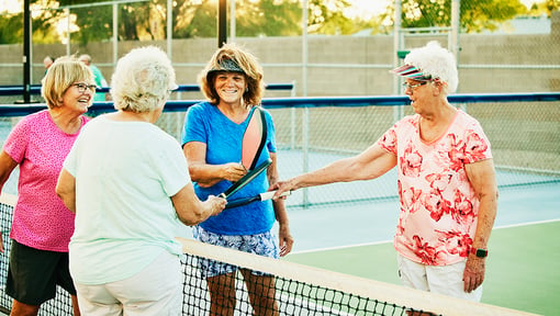 Four older adult pickleball players gathered at the net, tapping paddles after a match on an outdoor court.