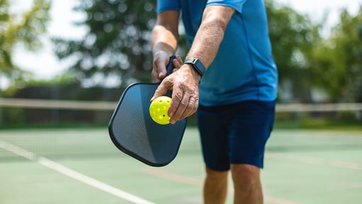 Close-up of a person holding a pickleball paddle and ball while standing on an outdoor pickleball court.