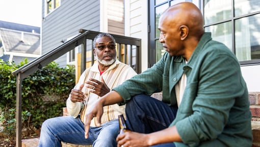 Two older adult men sitting on front steps and talking outside a residential building.