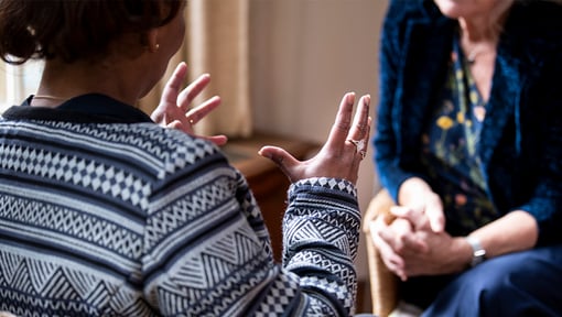 Two adults seated indoors having a face-to-face empathetic conversation, with one person gesturing while speaking.