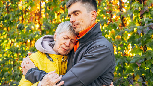 A man embracing an older woman in a leafy outdoor setting during daylight.